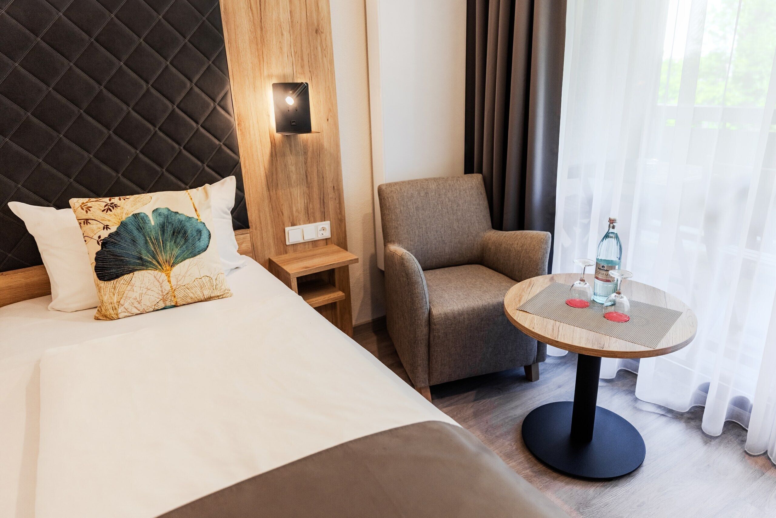 A hotel room with a bed, a chair, a small table with a bottle, and a wall-mounted light.