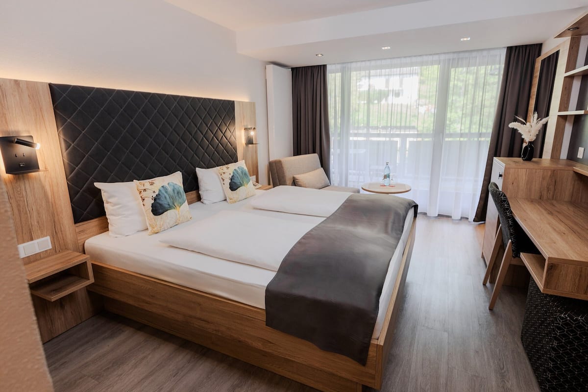 Une chambre d’hôtel moderne avec un grand lit, une tête de lit en bois, des tables de chevet, une petite table ronde et un bureau en bois.