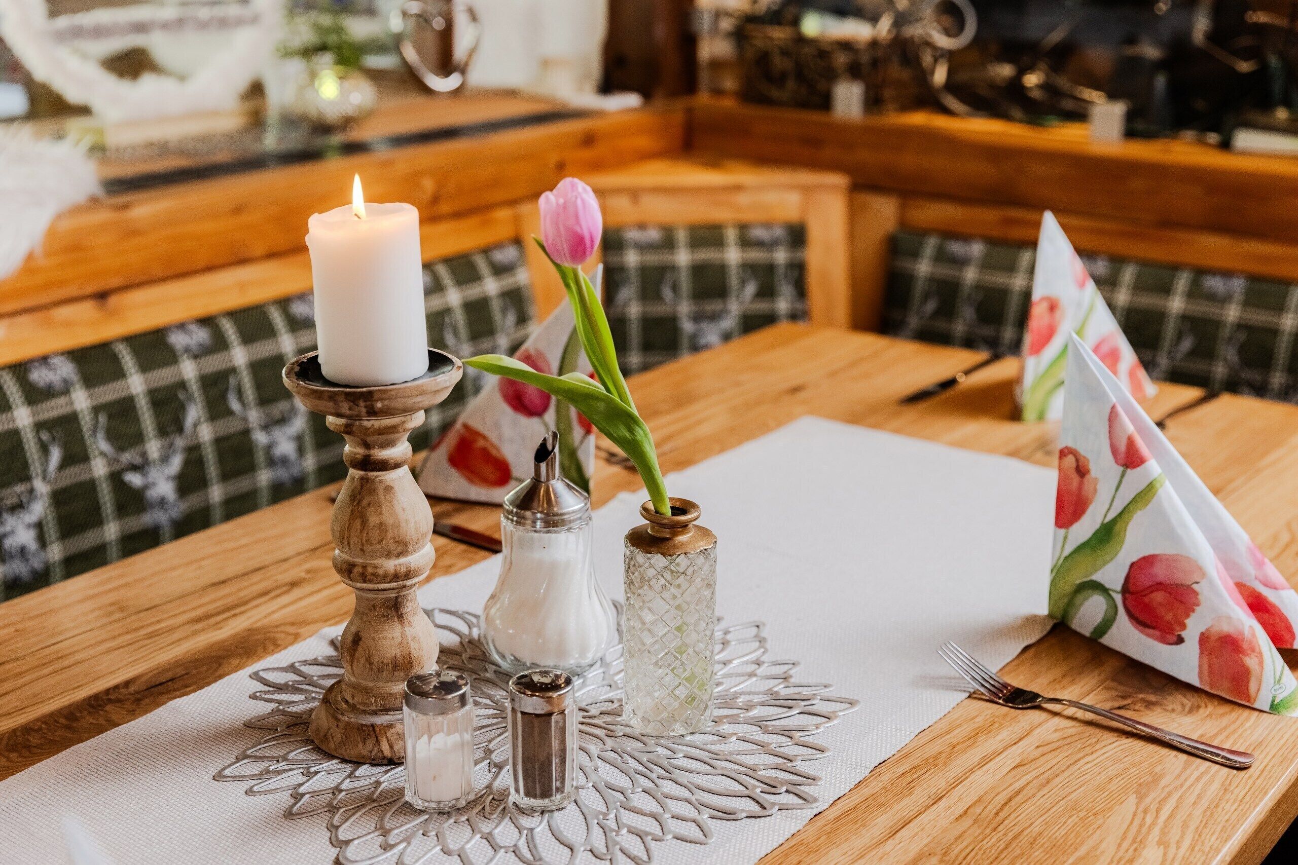 A wooden table with a white tablecloth, a lit candle, salt and pepper shakers, a glass vase with a tulip, and a napkin holder with a floral design.