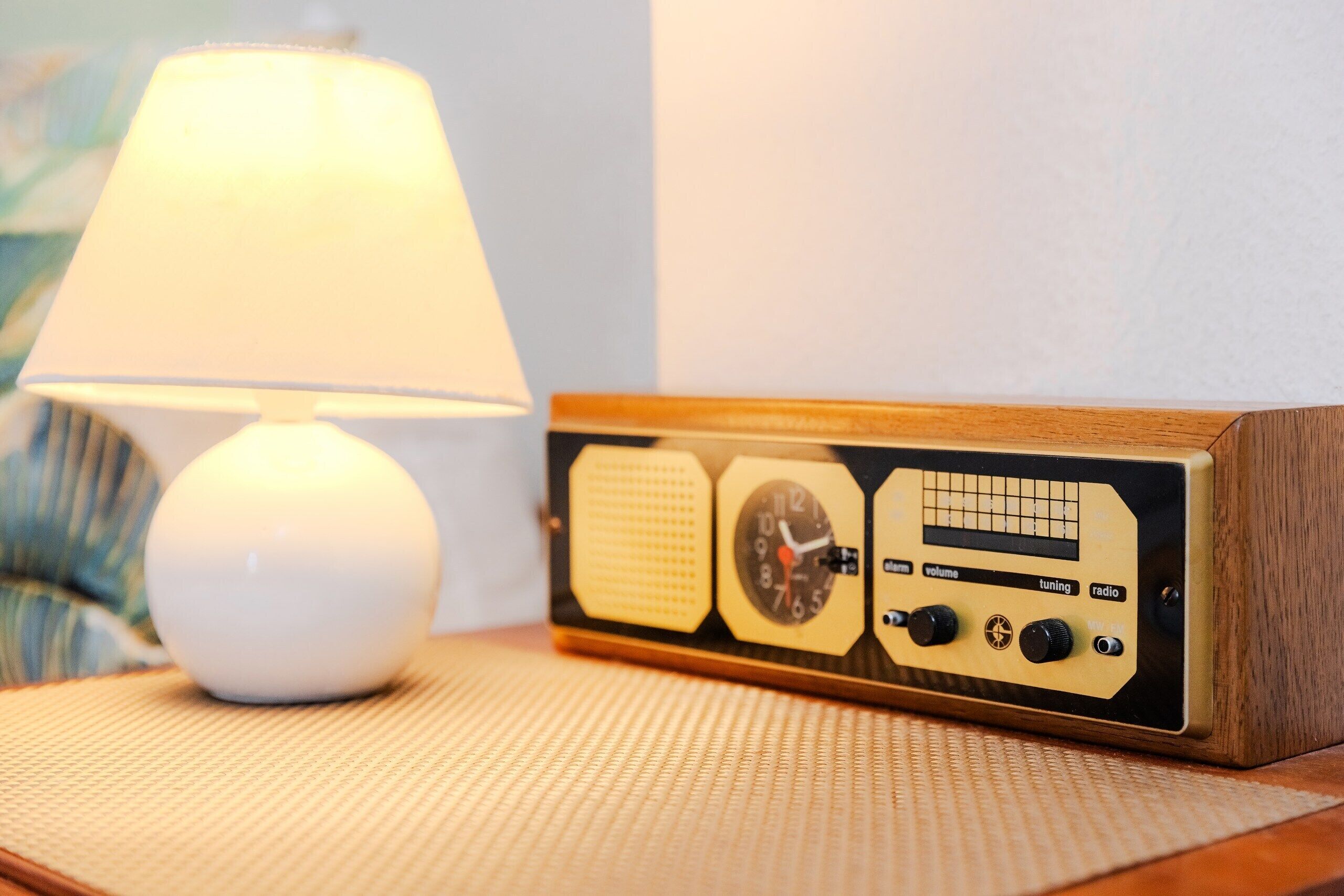 A radio with tuning controls and a clock, placed on a woven mat next to a lamp.