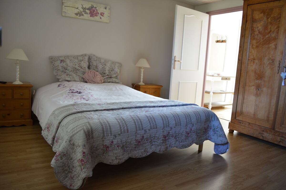 Un dormitorio con una cama, dos mesitas de noche, un armario de madera y una puerta que da a otra habitación.