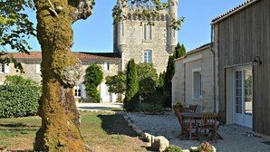 Un edificio de piedra con una torre, rodeado de un jardín con una mesa y sillas, y un árbol cubierto de musgo.