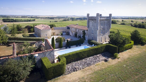 Un gran edificio de piedra con una torre, rodeado de jardines cuidados y una piscina.