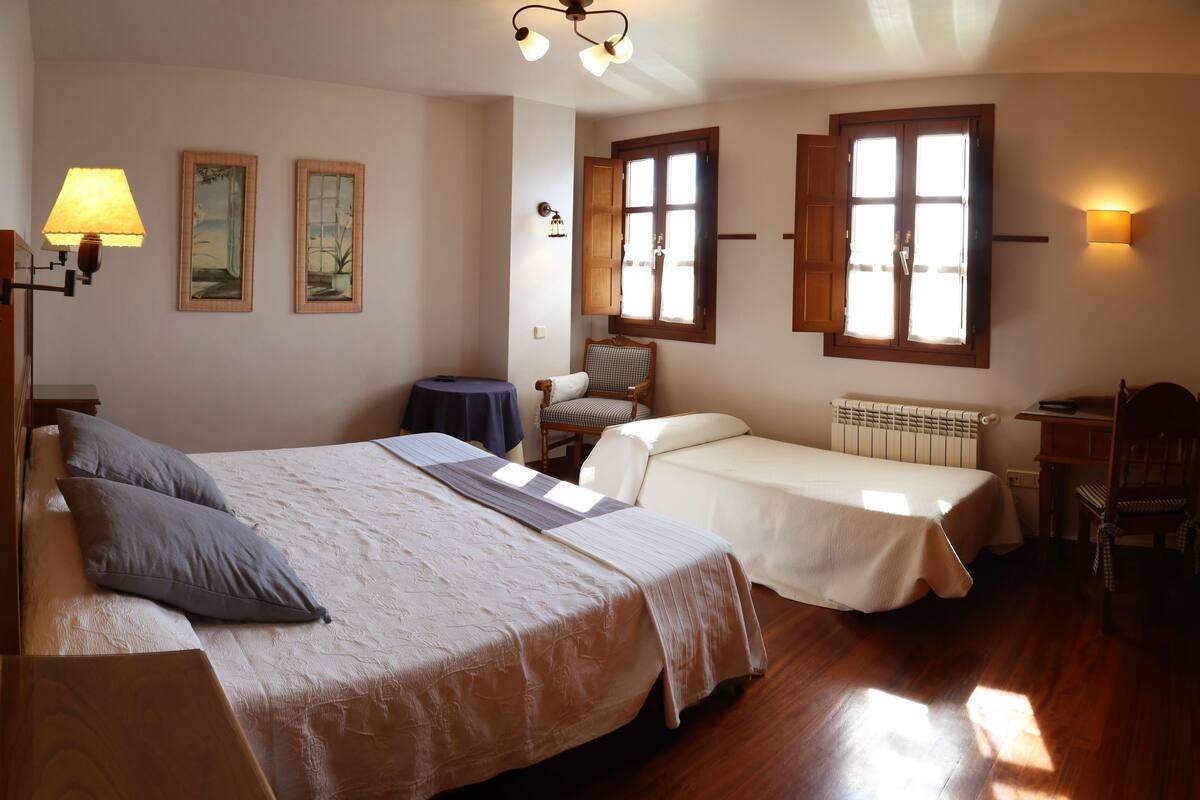Quarto com uma cama grande, uma cama menor, uma cadeira, uma mesa e um radiador.
