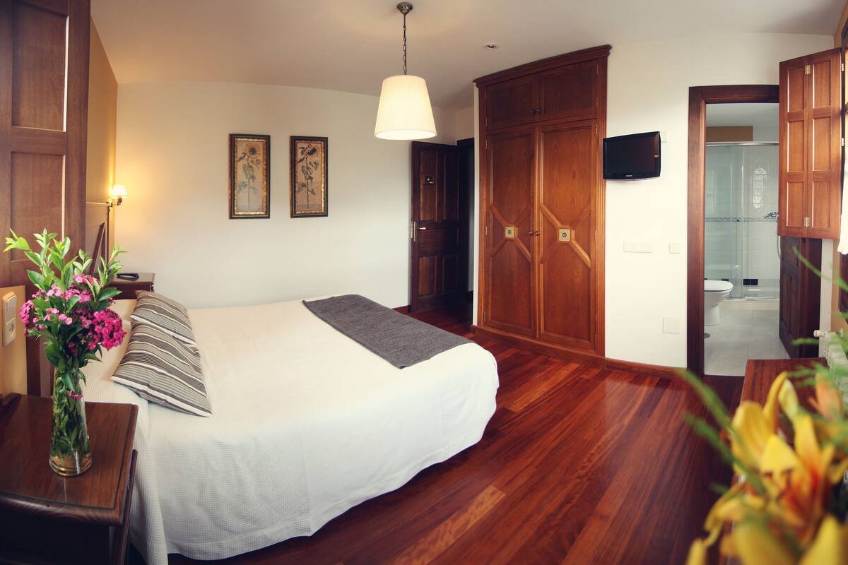 Quarto de hotel com uma cama, guarda-roupa de madeira, televisão fixada na parede e um banheiro visível através de uma porta aberta.