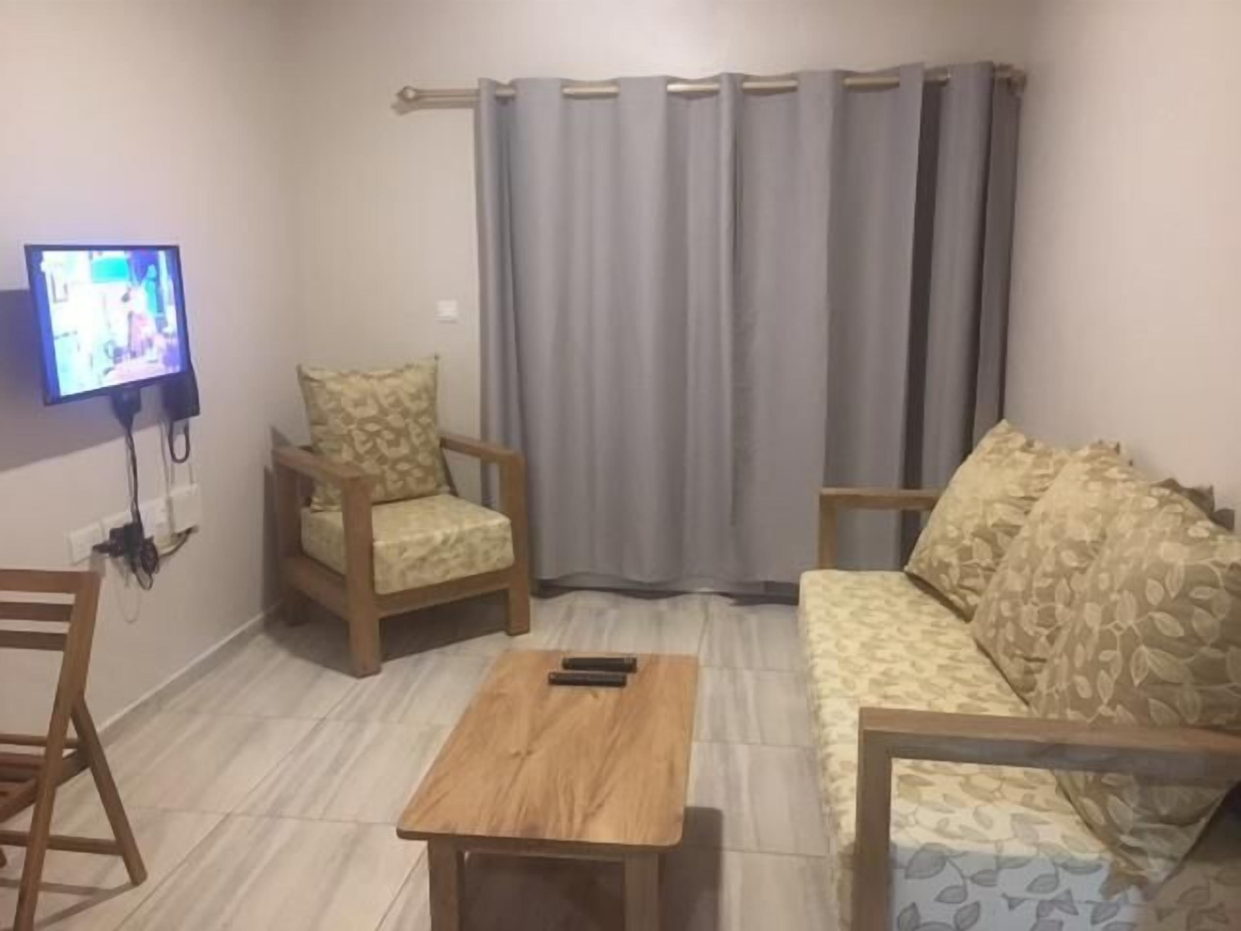 Un saló amb un sofà, dues butaques, una taula de centre, un televisor muntat a la paret i una finestra amb cortines.