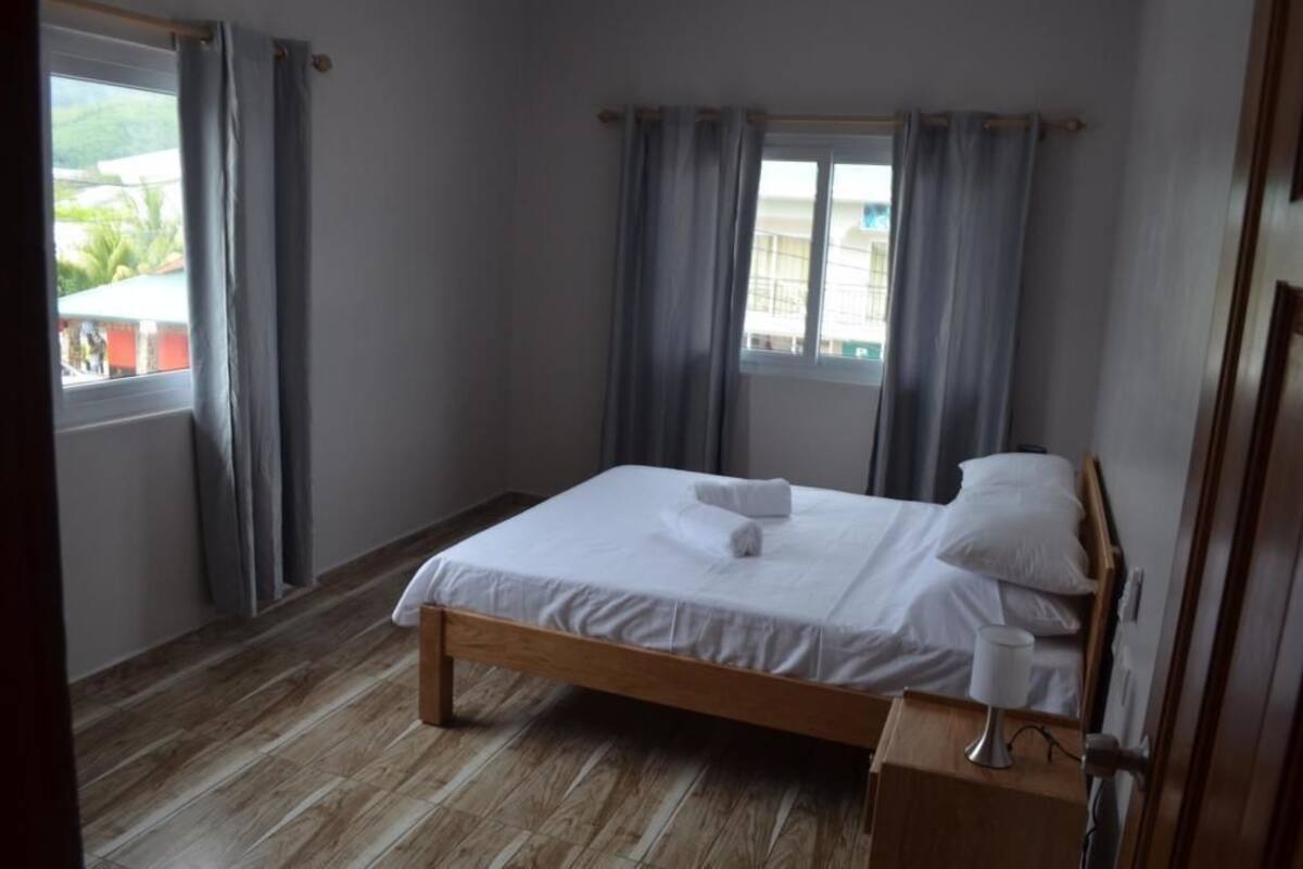 Un dormitorio con una cama de madera, ropa de cama blanca, una mesita de noche con lámpara y una ventana con cortinas.