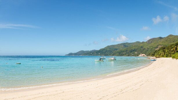 Una playa con agua turquesa cristalina, embarcaciones y una orilla arenosa con vegetación al fondo.