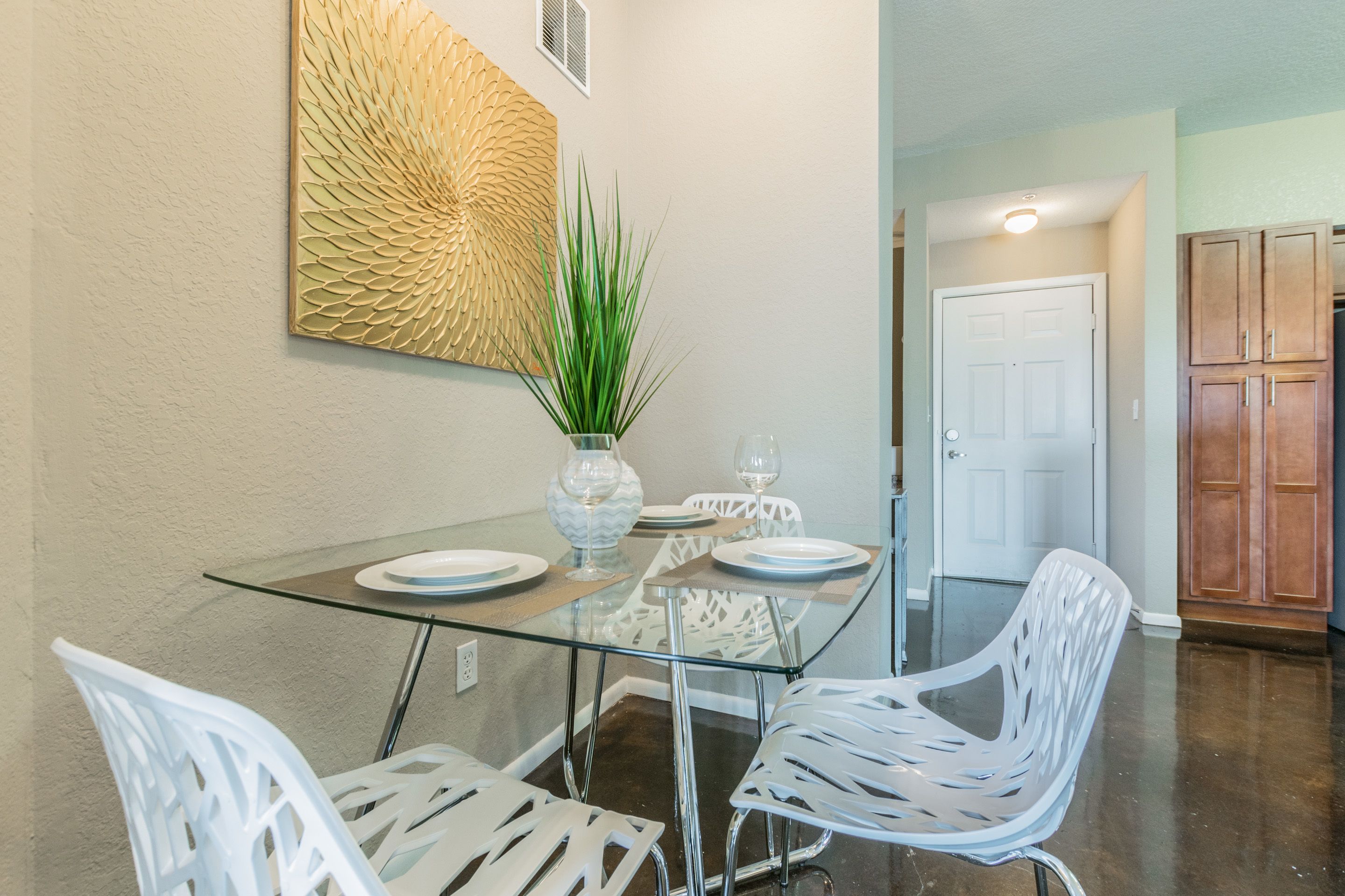 A dining area with a glass table, white chairs, a vase with a plant, and a decorative wall piece.
