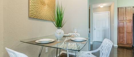 A dining area with a glass table, white chairs, a vase with a plant, and a decorative wall piece.