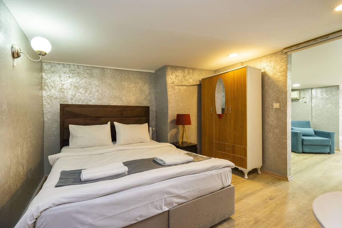 A neatly made bed with white linens, a wooden headboard, a wooden wardrobe, a red lamp, and a blue chair in the background.