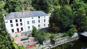 Un bâtiment blanc doté de nombreuses fenêtres et d’une porte rouge, entouré de verdure et bordé d’un petit plan d’eau.