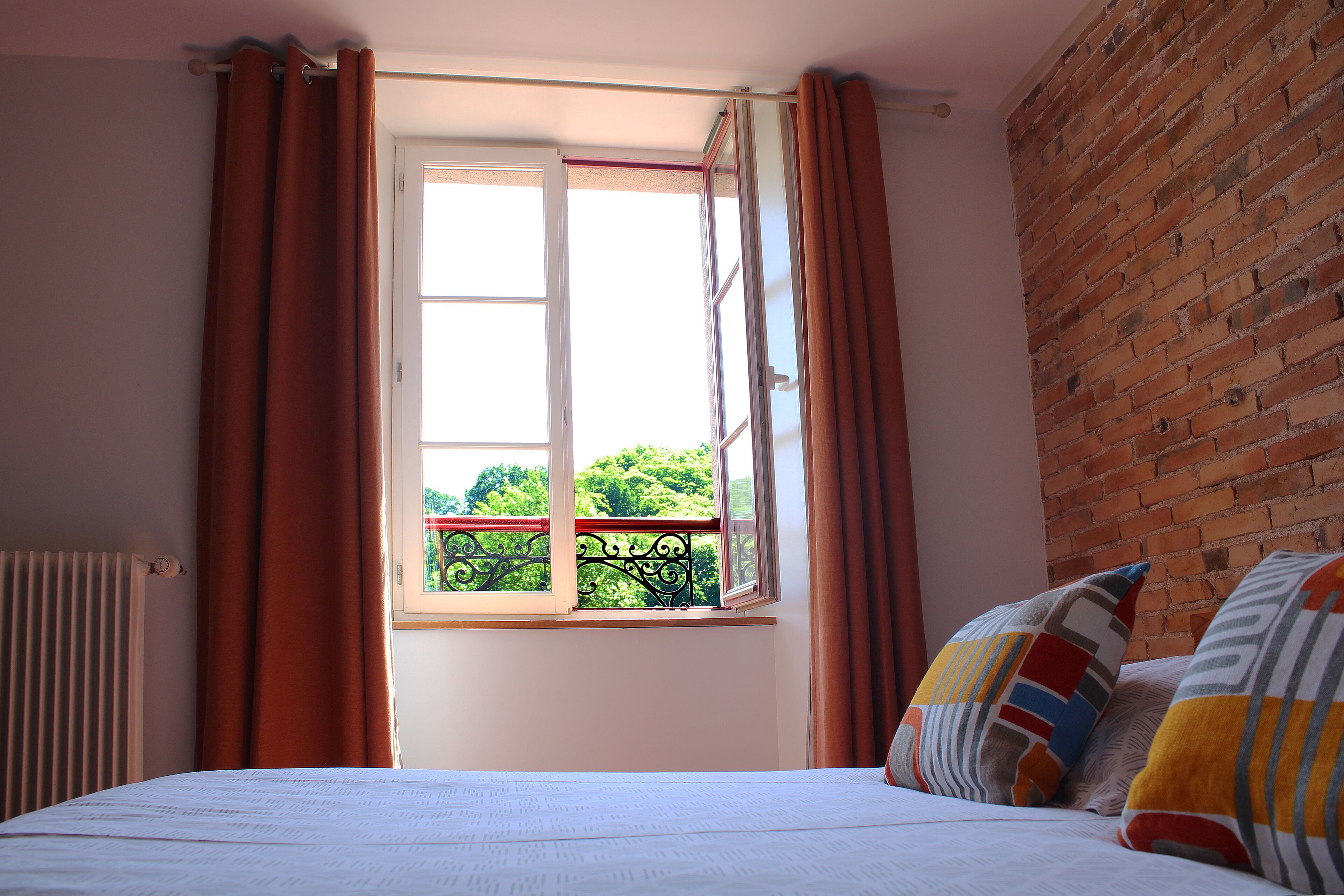 Een slaapkamer met een bed, een raam met gordijnen, een radiator en een bakstenen muur.