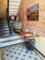Una escalera con barandilla de madera, un muro de ladrillo con un cuadro, una mesa con una botella y una máquina de coser.