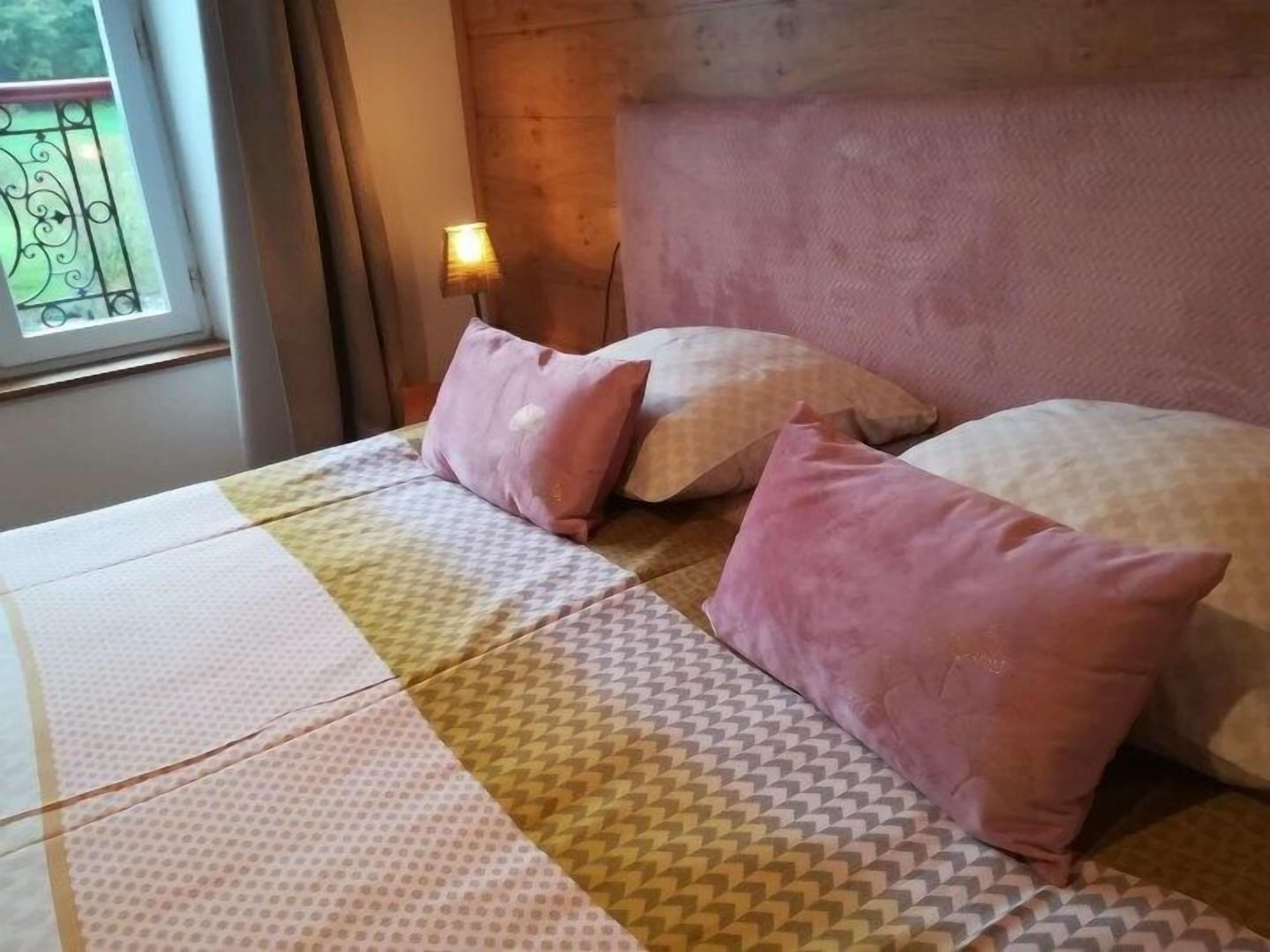 Uma cama com roupa de cama estampada e almofadas cor-de-rosa, uma mesa de cabeceira com abajur e uma janela com vista para áreas verdes.