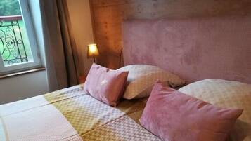 Uma cama com roupa de cama estampada e almofadas cor-de-rosa, uma mesa de cabeceira com abajur e uma janela com vista para áreas verdes.