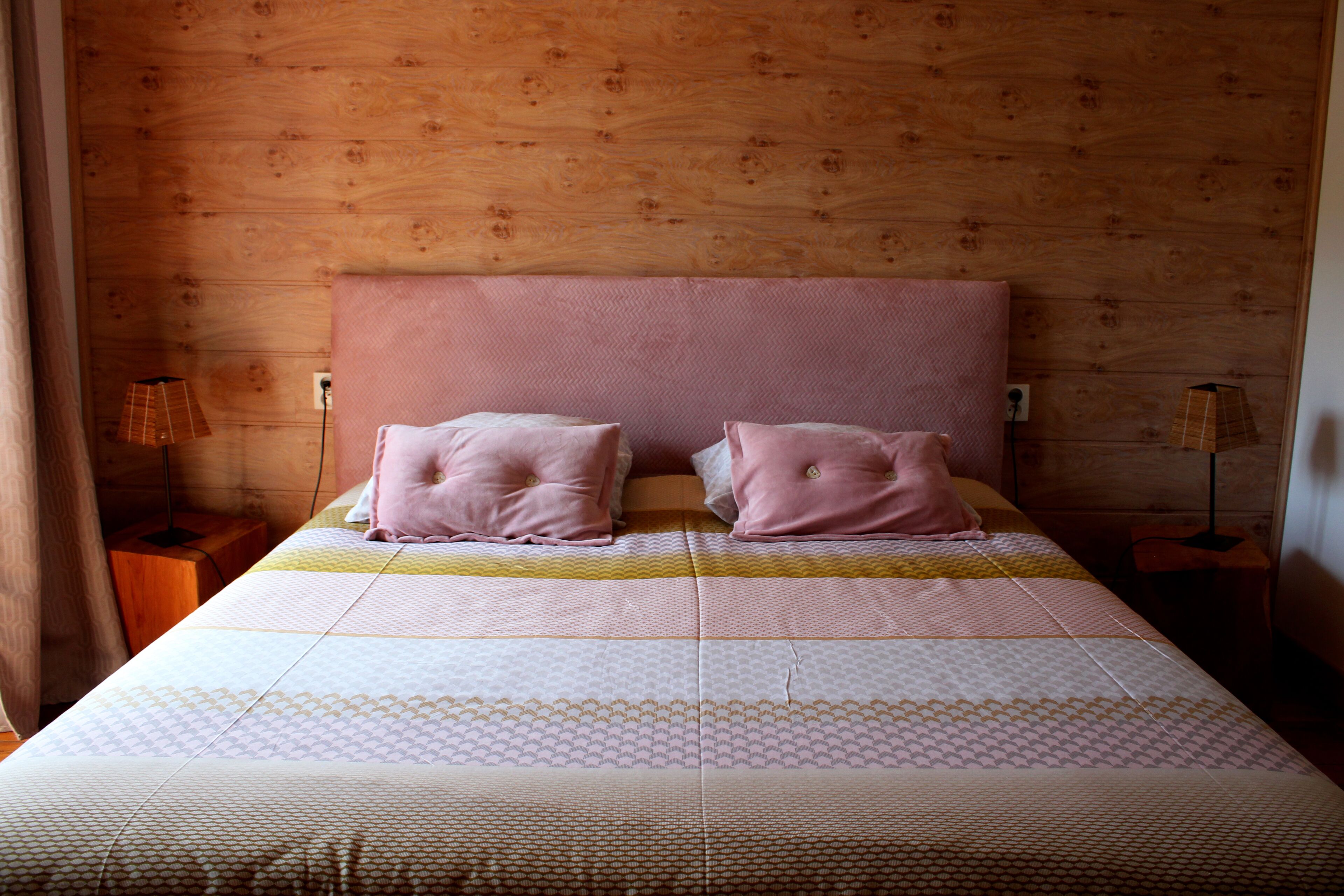 A bedroom with a wooden headboard, two pink pillows, a patterned bedspread, and a bedside table with a lamp.