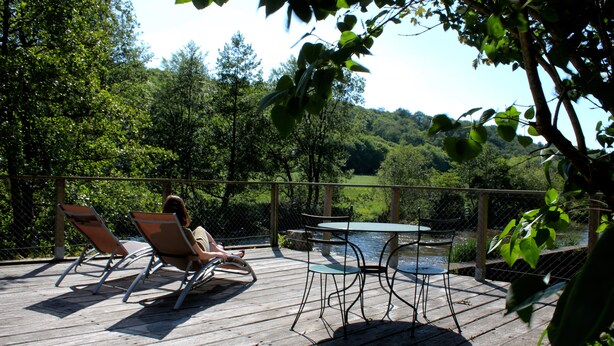 Une terrasse en bois, agréée de mobilier d’extérieur comprenant des chaises et une table, qui offre une vue sur une étendue d’eau et est entourée d’arbres.