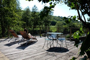 Terraza de madera con mobiliario exterior, incluyendo sillas y una mesa, con vistas a un cuerpo de agua rodeado de árboles.