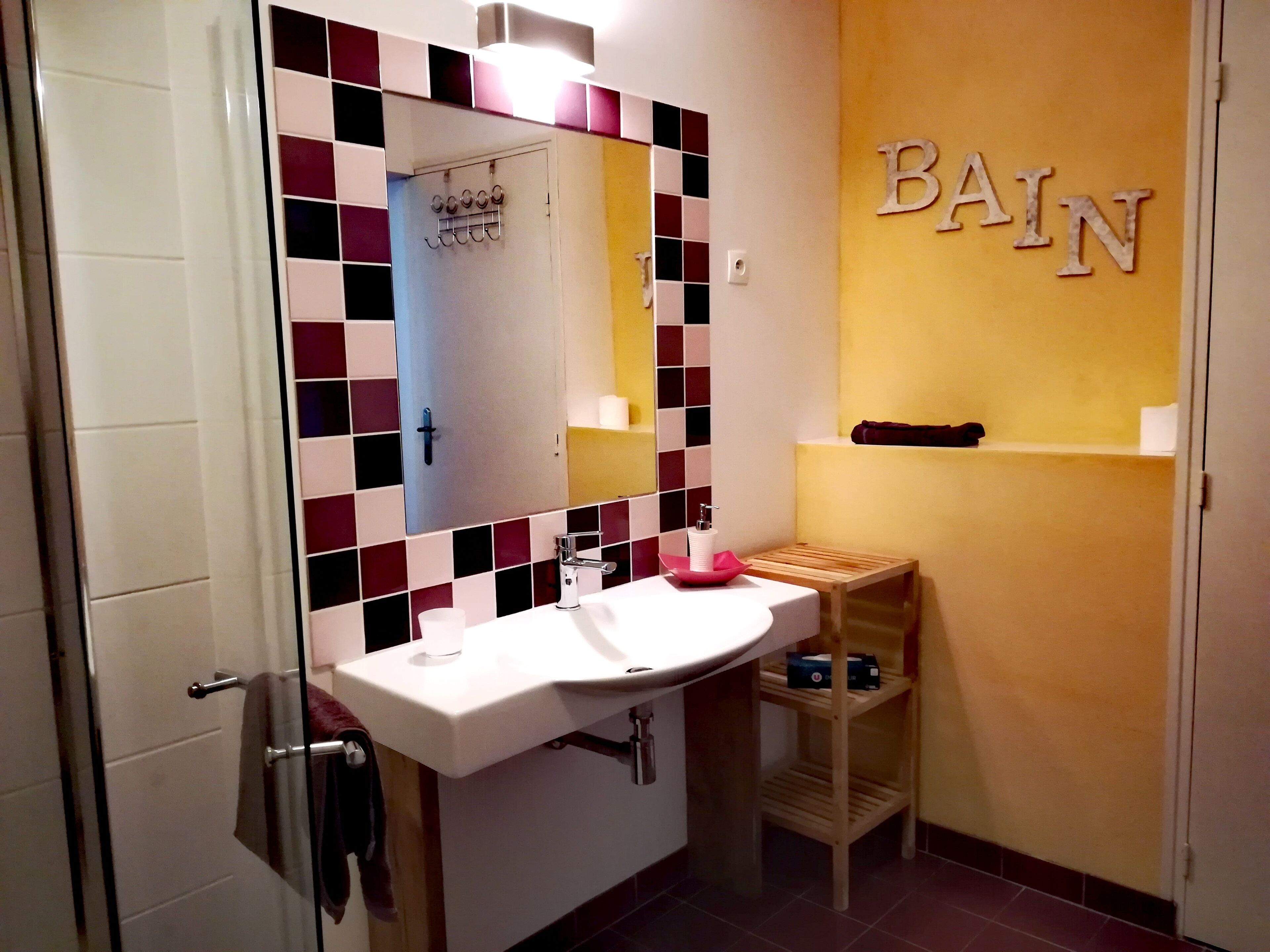 Een badkamer met een witte wastafel, een spiegel en een handdoekhouder. Op de muur hangt een bordje met de tekst 'BAIN' en een plank met toiletartikelen.
