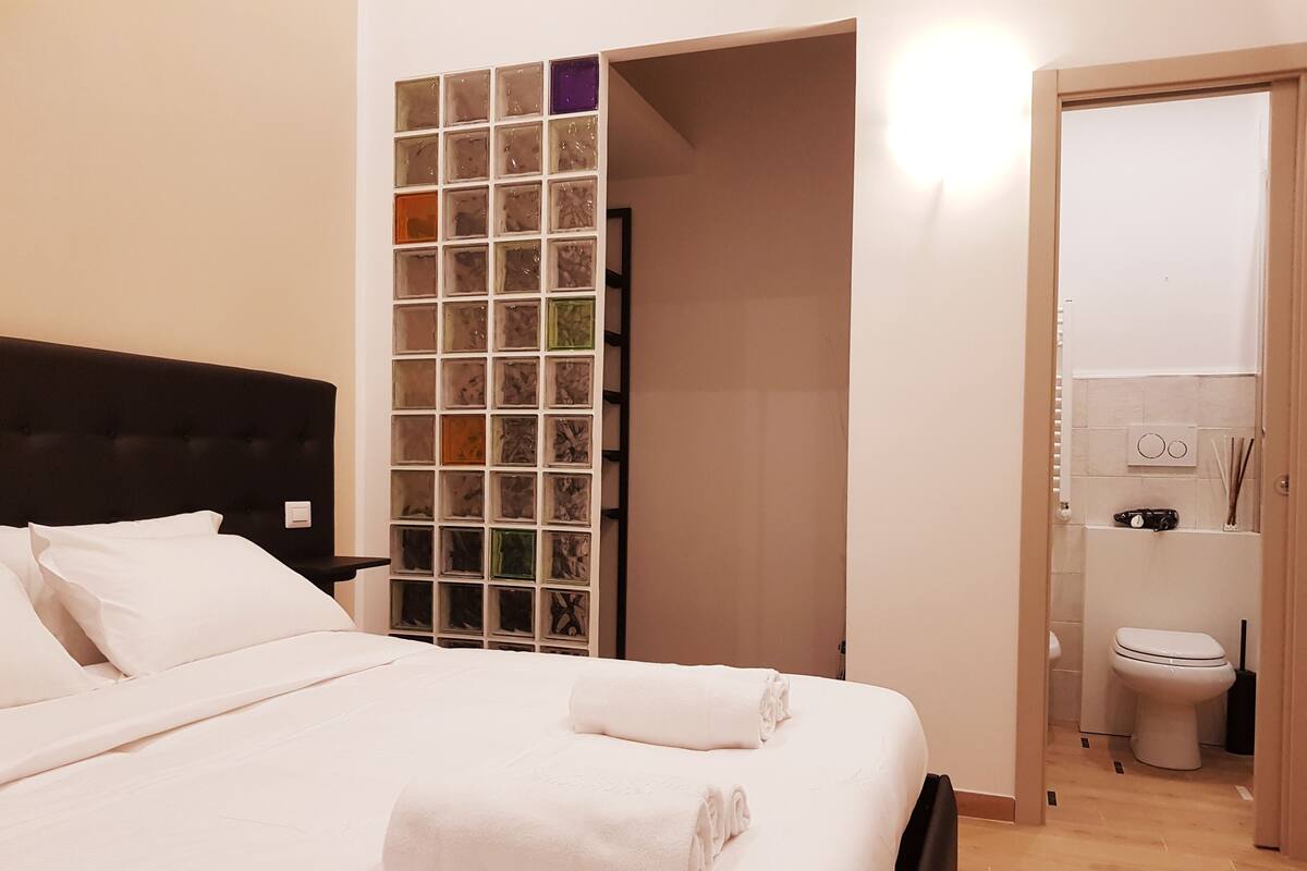 A neatly made bed with white linens and rolled towels, a black upholstered headboard, and a wall-mounted shelf with glass containers.