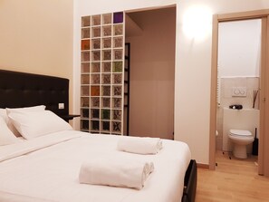 Una cama bien hecha con sábanas blancas y toallas enrolladas, un cabecero tapizado en negro y un estante montado en la pared con recipientes de vidrio.