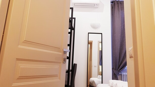 A narrow hallway leading to a bedroom with a bed, a chair, a mirror, and an air conditioning unit on the wall. - Cairoli Rooms (Rome)