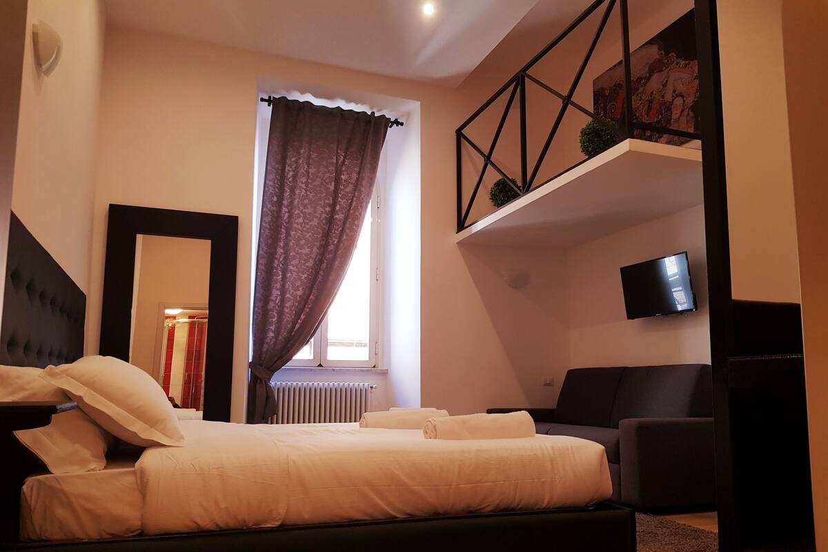 A modern bedroom with a large bed, a TV mounted on the wall, and a balcony with plants.
