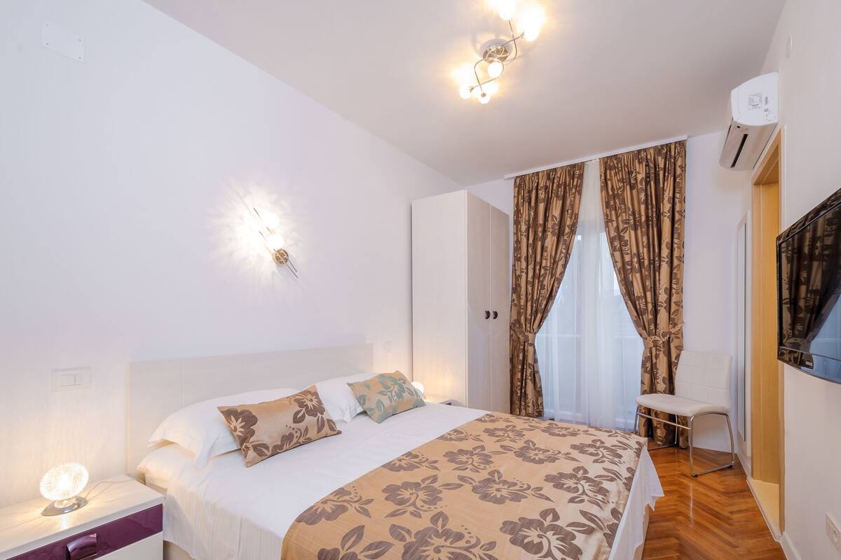 Ein Schlafzimmer mit Bett, Nachttisch, Lampe, Vorhängen und einem Fernseher.