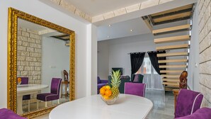 Un espace repas moderne avec une table blanche, des chaises violettes et un saladier rempli de fruits. Un grand miroir à cadre doré reflète la pièce.