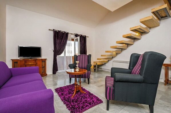 Una sala de estar con un sofá púrpura, sillones a rayas, un mueble de televisión de madera, una mesita redonda pequeña y una escalera con escalones de madera.