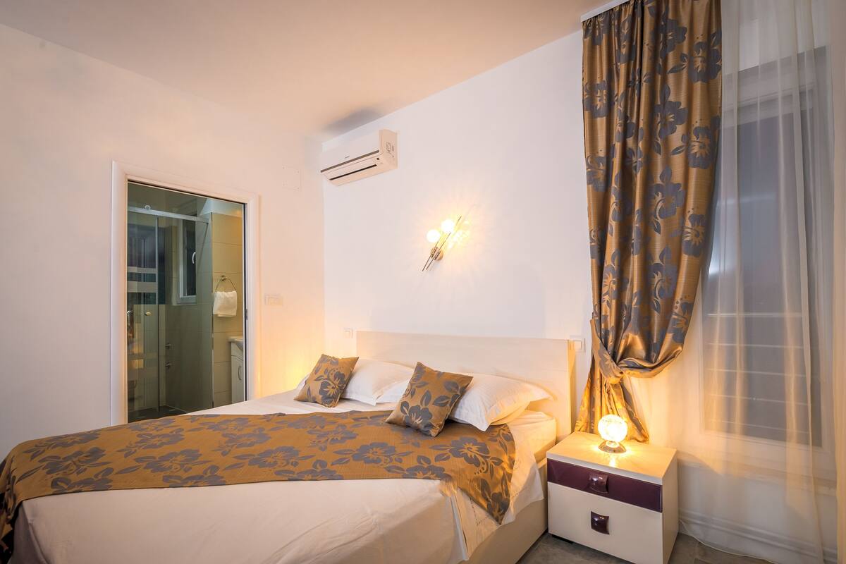 Ein Hotelzimmer mit Bett, Nachttisch, Wandlampe und einem Fenster mit Vorhängen.