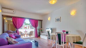 Un salon moderne avec un canapé violet, une table à manger avec des chaises et une grande fenêtre ornée de rideaux roses.