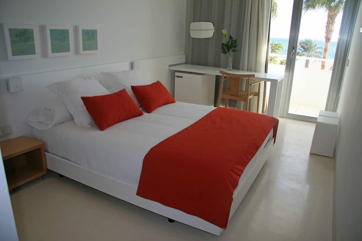 A bedroom with a bed, red pillows, a desk, and a view of the sea.
