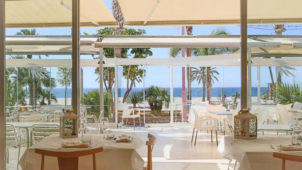 Un área de comedor al aire libre con mesas y sillas, ventanas amplias y vistas a la playa y el océano.