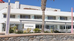 Un edificio moderno de dos plantas con fachada de vidrio, un restaurante visible llamado "Las Palmas" y mesas al aire libre con palmeras.
