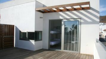 Un edificio moderno con terraza de madera, puerta de cristal que da a una habitación con cama y una pérgola de madera.