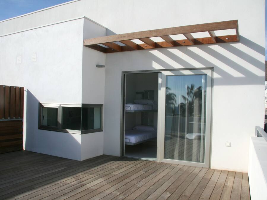 Un edificio moderno con terraza de madera, puerta de cristal que da a una habitación con cama y una pérgola de madera.