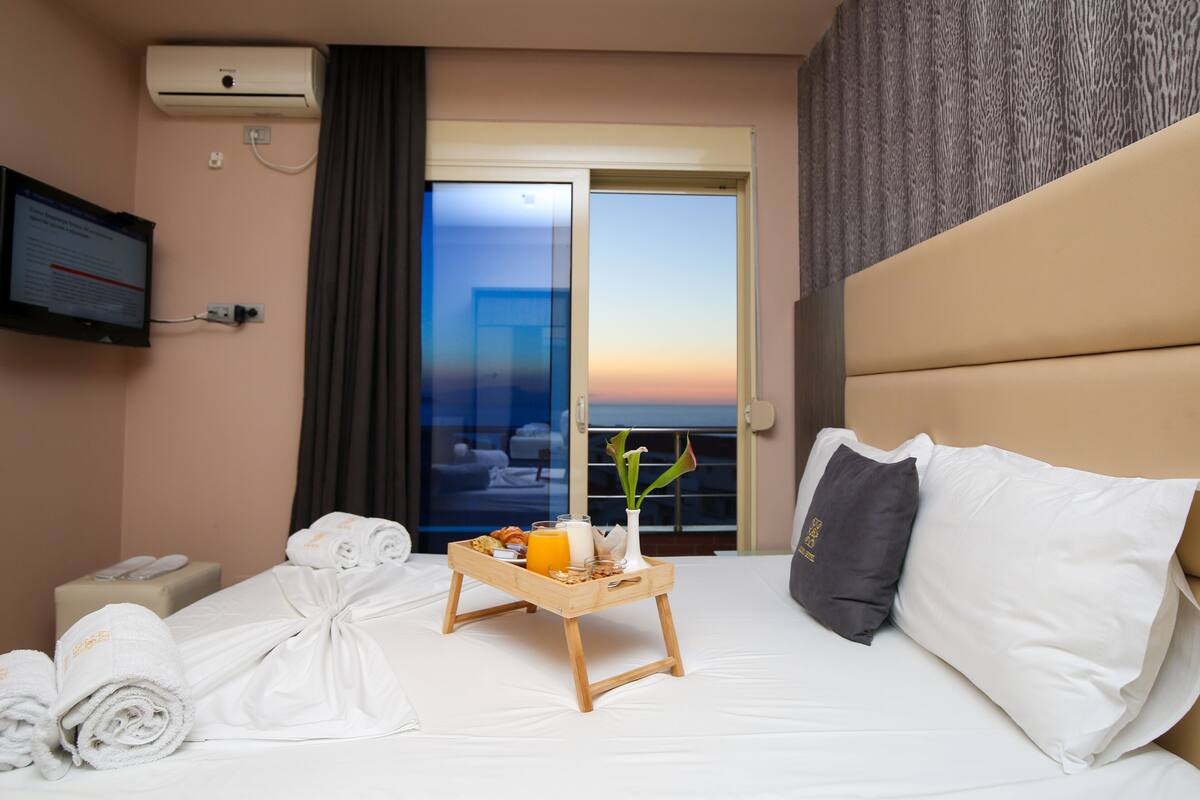 A hotel room with a bed, a bedside table with a tray of breakfast, a TV, and a view of the sunset through the window.