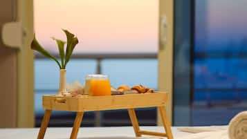 Un plateau en bois contenant des aliments pour le petit-déjeuner, posé sur un lit, un vase rempli de fleurs et une vue sur la mer au coucher du soleil.