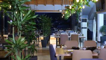Un comedor interior con mesas y sillas, plantas en macetas y luces colgantes.