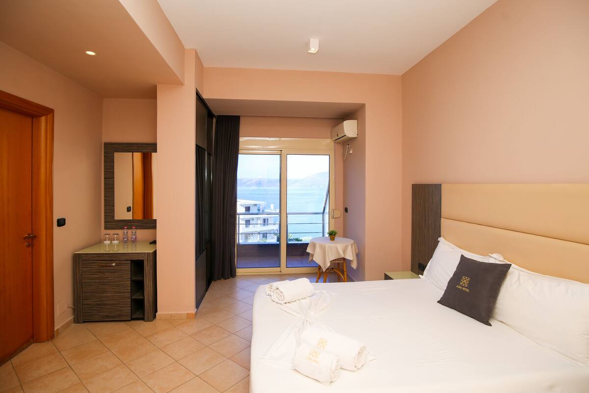 A hotel room with a bed, a desk, a chair, a small table, a mirror, and a view of the sea.
