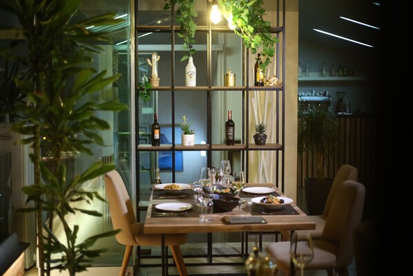 Comedor con separador de vidrio, mesa de madera, sillas y una variedad de plantas y botellas en estanterías.