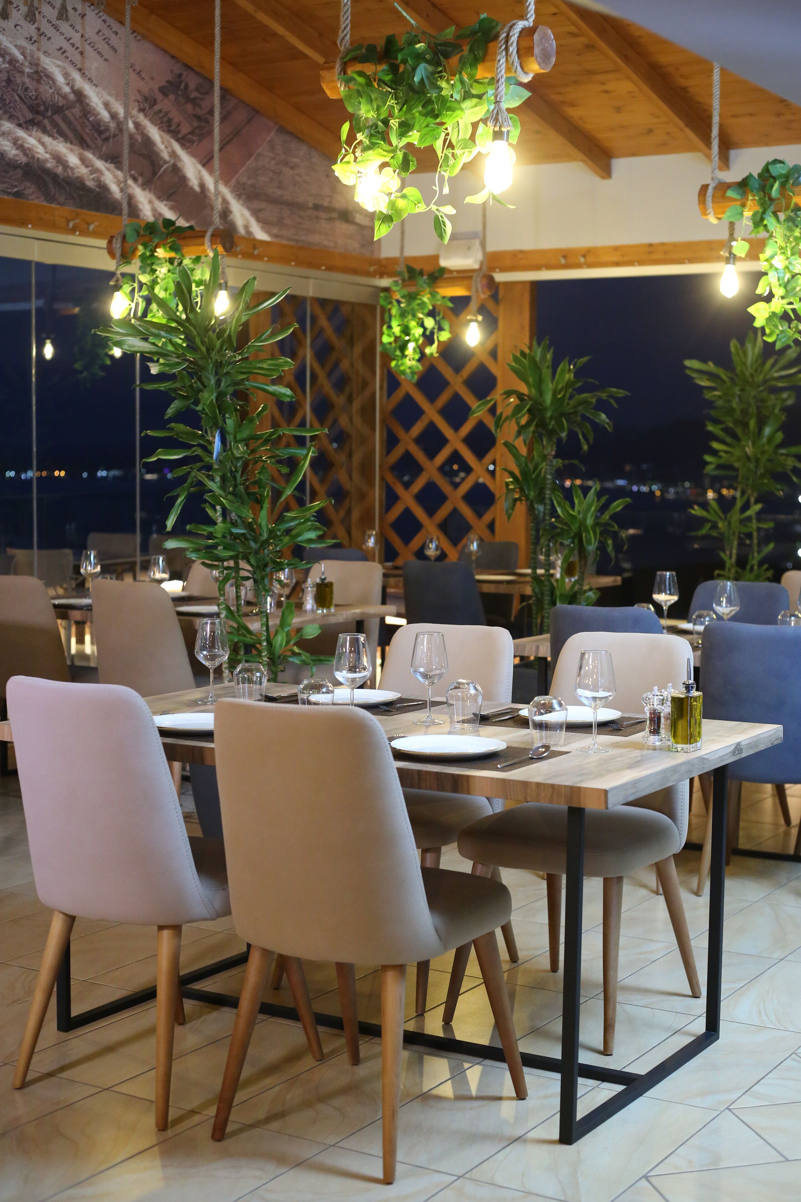 A restaurant with tables set for dining, chairs, hanging plants, and a city view.
