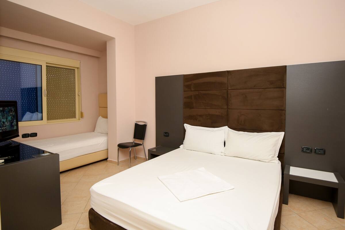 A hotel room with a large bed, a smaller sofa bed, a TV, a chair, and a desk.