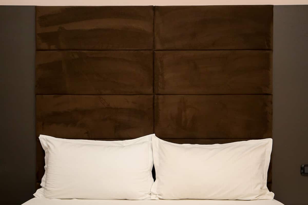 A neatly made bed with white linens and pillows, a wooden headboard, and a plain wall.