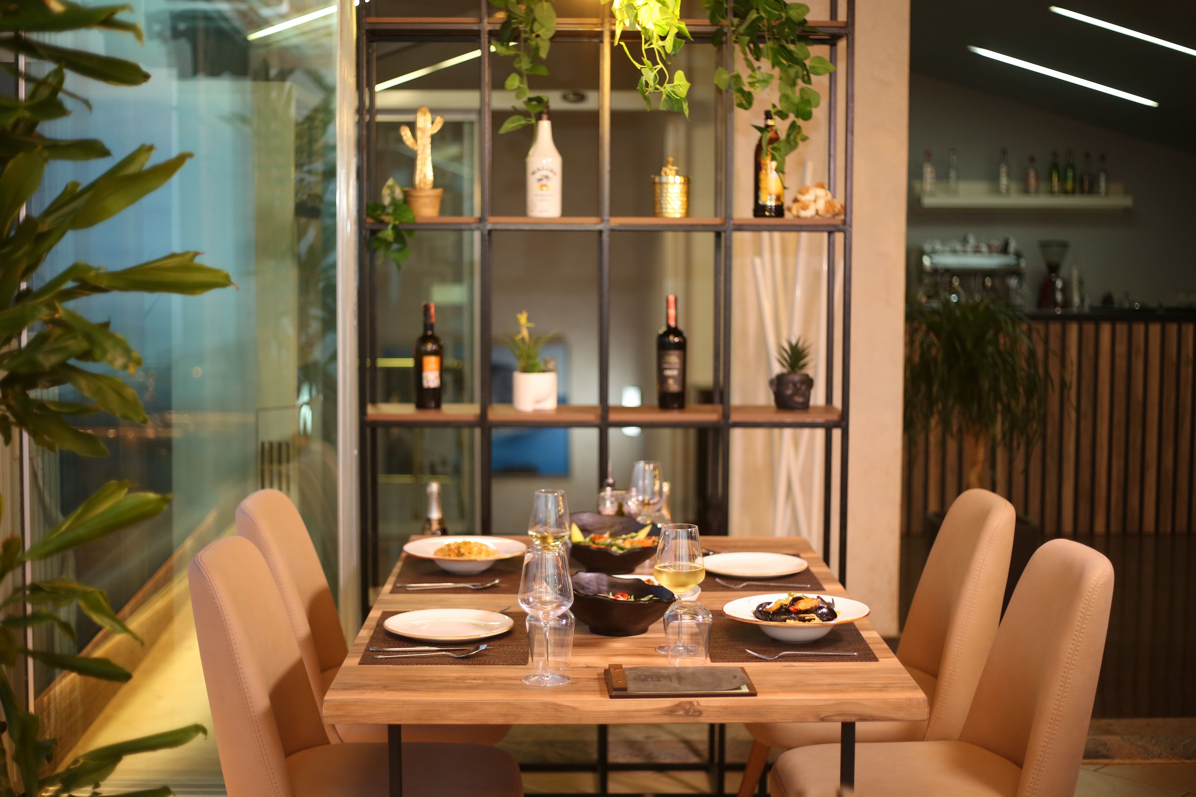 Un comedor con una mesa de madera dispuesta para cuatro personas, rodeada de sillas acolchadas. Un separador de vidrio divide el comedor de la zona del bar, que cuenta con estantes con botellas y plantas.
