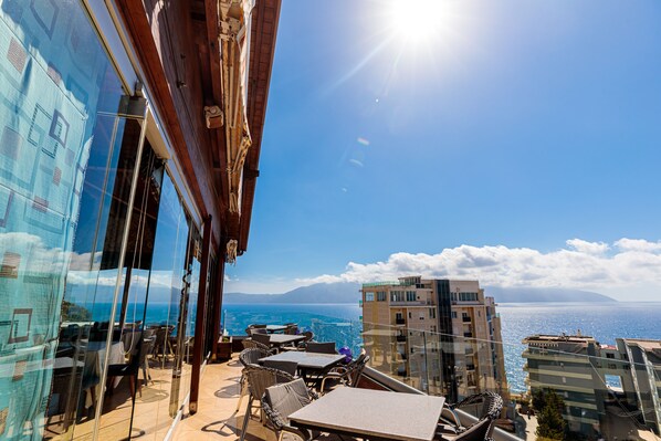 Una terraza con mesas y sillas que da al mar, un edificio de cristal a la izquierda y un cielo despejado.