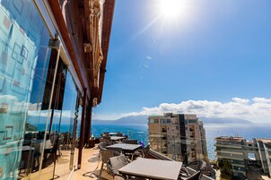 Una terraza con mesas y sillas que da al mar, un edificio de cristal a la izquierda y un cielo despejado.