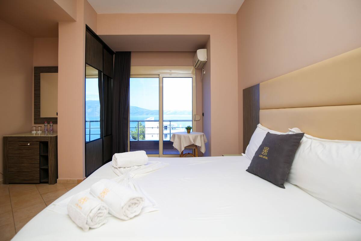 A hotel room with a large bed, a bedside table with a lamp, a small table with a plant, and a view of the sea through the window.